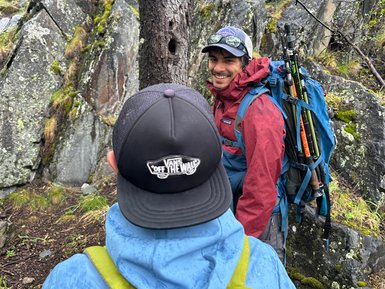 Zwei Wanderer stehen auf einem schmalen Pfad zwischen großen Felsen, umgeben von üppigem, grünem Laub.