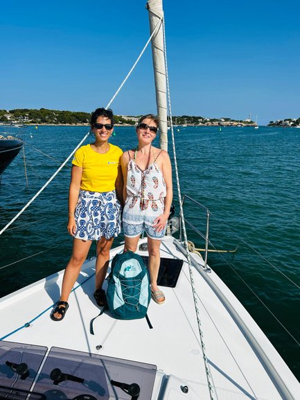 Zwei Frauen stehen auf dem Deck eines Segelbootes, umgeben von klarem Wasser und einem strahlend blauen Himmel.