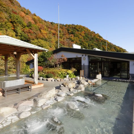 Ein entspannender Onsen-Bereich mit einem Holzpavillon und einem klaren Wasserbecken, umgeben von herbstlichen Bäumen.