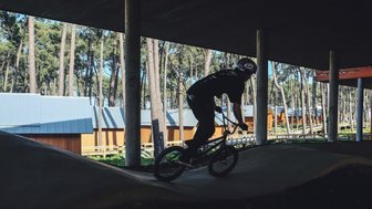 Ein BMX-Fahrer vollbringt eine beeindruckende Leistung auf einer kurvigen Strecke, umgeben von hohen Bäumen und modernen Hütten.
