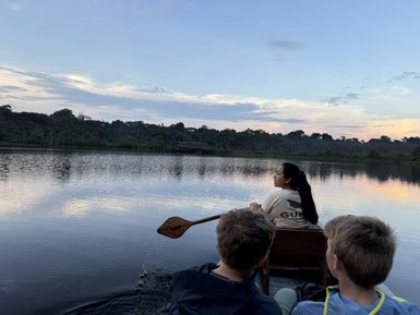 Eine Frau paddelt in einem Kanu auf einem ruhigen See, während zwei Jungen ihr aufmerksam zuschauen.
