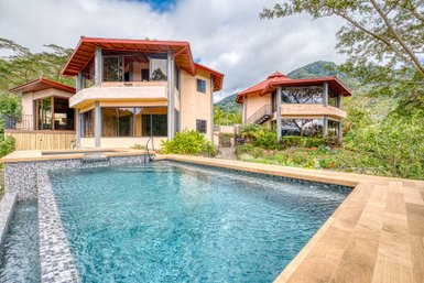 Zwei moderne Häuser mit roten Dächern stehen neben einem glitzernden Pool, umgeben von üppigem Grün und Bergen im Hintergrund.