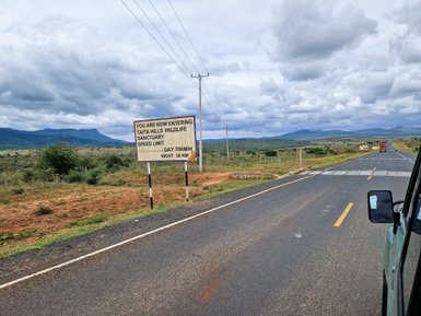 Ein Schild markiert den Eingang zum Taita Hills Wildlife Sanctuary, umgeben von sanften Hügeln und bewölktem Himmel.