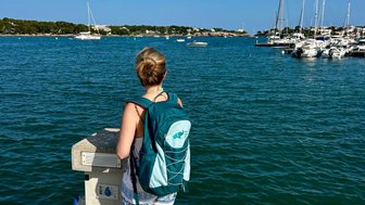 Eine Frau mit einem Rucksack steht am Hafen und blickt auf das ruhige Wasser, umgeben von Segelbooten und einer klaren blauen Himmel.