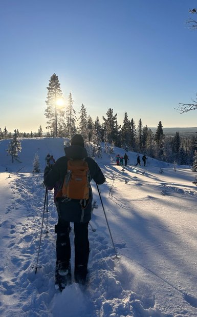 Ein Wanderer mit einem orangefarbenen Rucksack geht durch den tiefen Schnee, während die Sonne hinter den Bäumen aufgeht.