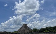 Eine beeindruckende Pyramide erhebt sich vor einem strahlend blauen Himmel mit flauschigen Wolken, umgeben von üppigem Grün.
