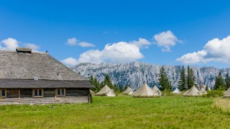 Eine rustikale Holzhütte steht im Vordergrund, umgeben von Zeltlagern und majestätischen Bergen unter einem strahlend blauen Himmel.