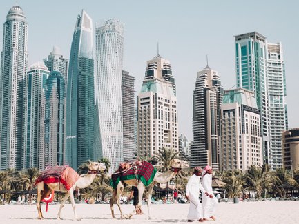 Zwei Kamele mit bunten Decken stehen am Strand von Dubai, während zwei Männer in traditioneller Kleidung vorbeigehen.