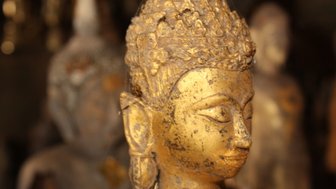 Eine goldene Buddha-Statue mit feinen Details und einem ruhigen Ausdruck, umgeben von weiteren, unscharfen Statuen im Hintergrund.