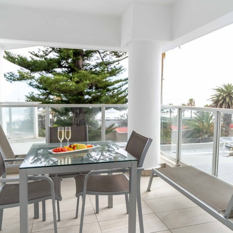 Ein eleganter Balkon mit einem Glastisch, zwei Sektgläsern und frischem Obst, umgeben von Palmen und Meerblick.