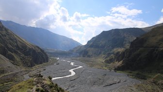 Ein malerisches Tal im Kaukasus mit einem geschlängelten Fluss, umgeben von majestätischen Bergen und bewölktem Himmel.