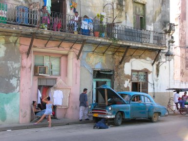 Eine lebhafte Straßenszene in Havanna mit einem blauen Oldtimer, der repariert wird, während Menschen im Hintergrund entspannen.