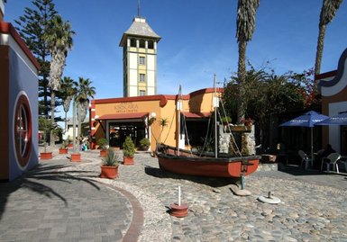 Ein malerischer Platz in Swakopmund mit einem historischen Gebäude und einem Boot, umgeben von Palmen und bunten Blumen.