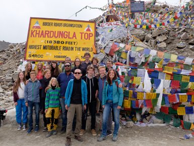 Eine Gruppe von Reisenden posiert fröhlich vor dem Khardungla-Pass-Schild, umgeben von bunten Gebetsfahnen.