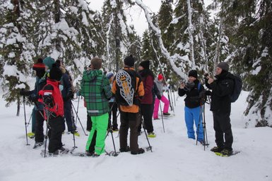 Eine Gruppe von Menschen in bunten Winterkleidung steht im Schnee und bereitet sich auf eine Schneeschuhwanderung vor.