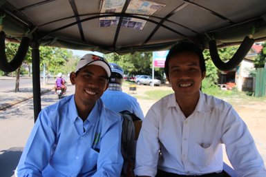 Zwei Männer lächeln fröhlich in einem Tuk-Tuk, während sie durch eine belebte Straße fahren, umgeben von Bäumen und Fahrzeugen.