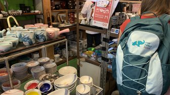 Eine Person mit einem Rucksack steht in einem Geschäft voller bunter Geschirrteile und Töpfe auf Regalen.