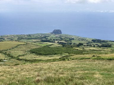 Eine weite Landschaft mit sanften Hügeln, üppigem Grün und einem Blick auf das blaue Meer im Hintergrund.