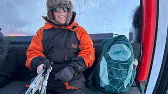 Eine Person in einem orange-schwarzen Schneeanzug sitzt in einer Gondel, umgeben von frostigen Fenstern und einem Rucksack.