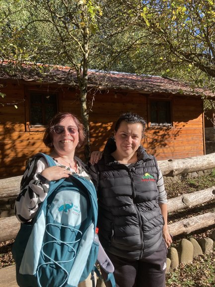Zwei Frauen stehen lächelnd vor einer Holzlaube, umgeben von Bäumen und einem sonnigen, klaren Himmel.