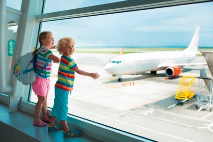 Zwei fröhliche Kinder stehen am Fenster eines Flughafens und beobachten ein Flugzeug auf dem Rollfeld.