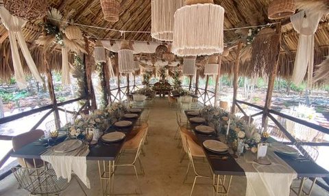 Ein stilvoll dekorierter Essbereich mit langen Tischen, umgeben von Pflanzen und einem rustikalen Strohdach.