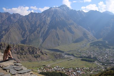 Eine Person sitzt am Rand einer Klippe und blickt auf eine weite, grüne Tallandschaft mit Bergen im Hintergrund.