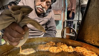 Ein älterer Mann bereitet frisch frittierte Süßigkeiten in einer großen Pfanne auf einem belebten Markt in Delhi zu.