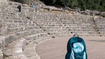 Eine Gruppe von Menschen erkundet die steinernen Stufen eines antiken Theaters, während ein Rucksack im Vordergrund steht.