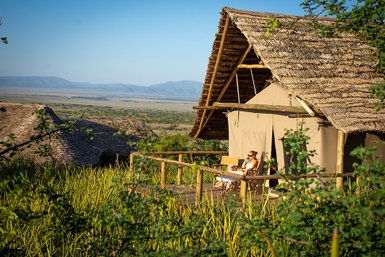 Eine Frau sitzt entspannt auf der Veranda eines Zeltes und genießt die Aussicht auf die weite Landschaft des Ngorongoro-Kraters.