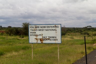 Ein rostiger Schild zeigt den Eingang zum Tsavo West Nationalpark mit Geschwindigkeitsbegrenzungen für Tag und Nacht.