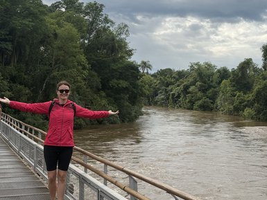 Eine Frau in einem pinken Kleid steht auf einer Holzplanke und breitet die Arme aus, um den malerischen Fluss zu genießen.