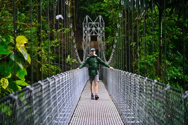 Ein Wanderer in einem lebhaften grünen Mantel steht gelassen auf einer Hängebrücke, umgeben von üppiger tropischer Vegetation.