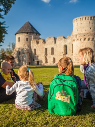 Eine Familie sitzt auf dem Gras vor einer alten Burg, während sie sich unterhält und die Sonne genießt.