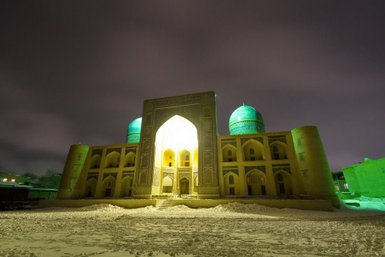 Die Mir-i-Arab-Medrese strahlt in der Nacht, beleuchtet von warmem Licht, umgeben von schneebedecktem Boden.