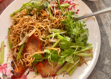 Ein Teller mit gebratenen Nudeln, frischen Kräutern und gegrilltem Fleisch, umgeben von verschiedenen Beilagen.