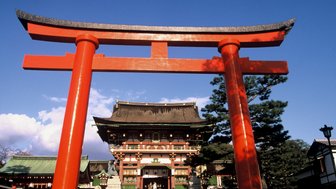 Ein leuchtend rotes Torii steht majestätisch vor einem traditionellen japanischen Schrein unter einem klaren blauen Himmel.