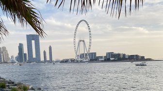 Ein Blick auf das Riesenrad, umgeben von modernen Gebäuden und dem ruhigen Wasser des Hafens, unter einem bewölkten Himmel.