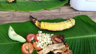 Auf einem großen Blatt liegen gegrillte Bananen, frisches Gemüse und zarte Fleischstücke, die ein einladendes Picknick bilden.