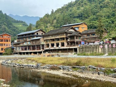 Traditionelle Holzhäuser stehen am Ufer eines ruhigen Flusses, umgeben von grünen Bergen und sanften Hügeln.