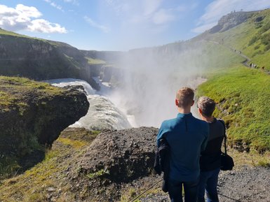 Zwei Personen stehen am Rand des Gullfoss-Wasserfalls und beobachten die tosenden Wassermassen, umgeben von grüner Landschaft.