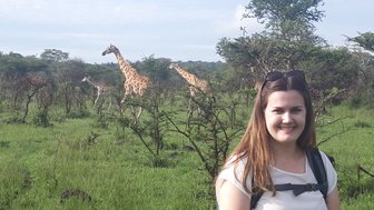 Eine Frau mit Rucksack lächelt vor einer Gruppe von Giraffen, die in der grünen Savanne von Mburo Nationalpark grasen.