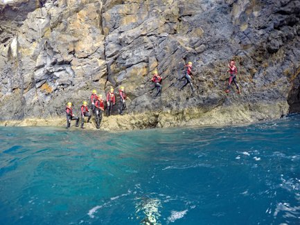 Eine Gruppe von Abenteurern in Neoprenanzügen klettert an einer steilen Felswand entlang, umgeben von klarem, blauem Wasser.