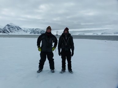 Zwei Männer stehen auf einer schneebedeckten Fläche, umgeben von Bergen und einem Fjord im Hintergrund.