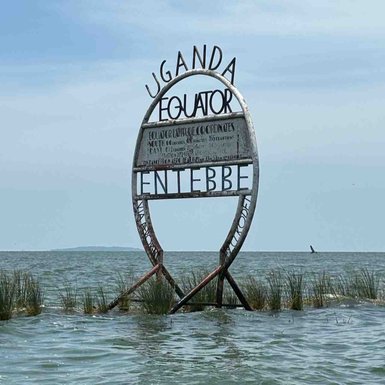 Ein großes, metallisches Schild markiert den Äquator in Uganda, umgeben von Wasser und Gras im Hintergrund.