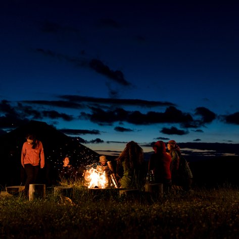 Eine Gruppe von Menschen versammelt sich um ein Lagerfeuer, während der Himmel in der Dämmerung leuchtet.