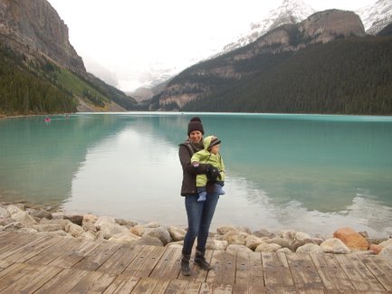 Eine Frau steht am Ufer eines klaren Sees in Kanada und hält ein Baby in einem grünen Anzug in ihren Armen.