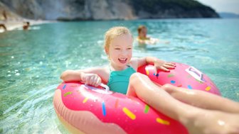 Ein fröhliches Mädchen mit blonden Locken genießt das Wasser in einem bunten Donut-Floatie an einem sonnigen Strand.