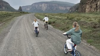 Eine Familie fährt mit Fahrrädern auf einem unbefestigten Weg durch eine grüne Landschaft unter einem bewölkten Himmel.