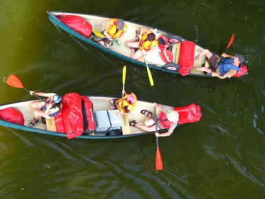 Zwei Kanus mit mehreren Personen paddeln auf einem ruhigen, grünen Gewässer, umgeben von einer friedlichen Naturkulisse.